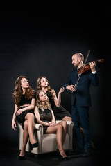 Three beautiful girls in black evening dresses are sitting in the chair, and the man plays the violin