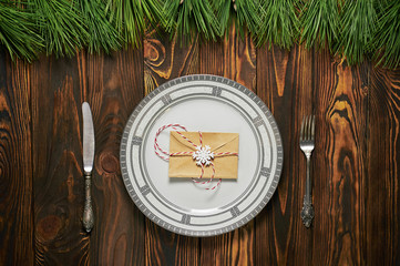 christmas table setting at brown wooden background with white plate, silver cutlery and paper envelope.