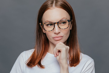 Thoughtful Caucasian woman with full lips, dark brown hair, looks thoughtfully aside, dressed in white clothes, being deep in thoughts, tries to make decision, poses over grey wall. Facial expressions