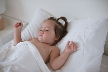 A little pretty dark-haired toddler girl is lying in bed smiling and looking happy.