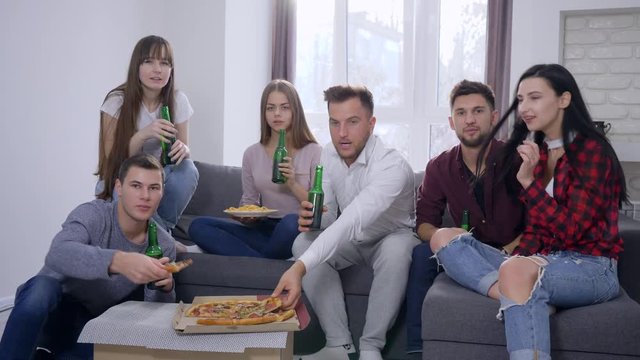 sport fans watching decisive moment of game and happy for victory sitting on the sofa in front of the TV eating pizza and making toast with glass bottles at home party.