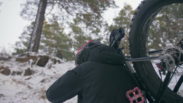 Professional extreme sportsman biker to bear fat bike to up mountain in outdoors. Cyclist walk in winter snow forest. Man walking with mountain bicycle with big tire in helmet, glasses. Slomo 60fps.