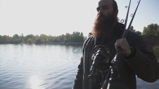 Bearded fisherman with long beard walks on the river bank with fishing rods. Slow motion.