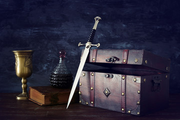 low key image of open chest, wine cup, antique old book and sword. fantasy medieval period.