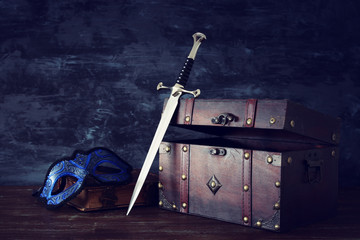 low key image of open chest, mysterious mask over antique old book and sword. fantasy medieval period.