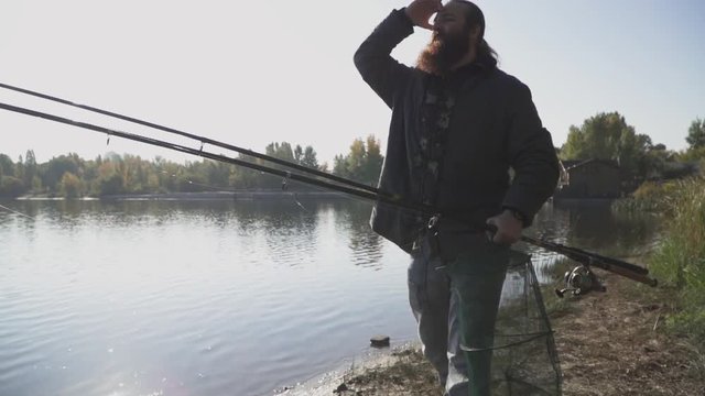 Lonely fisherman with long beard walks on the river bank with fishing rods. Slow motion.