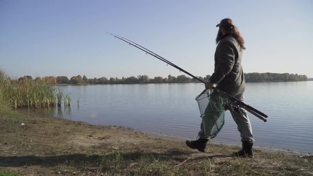 Fisherman with long beard walks near the river with fishing rods and ambush hoop net. Slow motion.