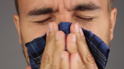 man with beard and mustache sneezes then blows his nose and wipes nose with a blue handkerchief, face closeup, gray background, front view
