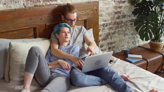 Handheld shot of cheerful young woman with blue hair relaxing on bed and chatting with bearded man in glasses working on laptop