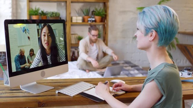 Medium shot of young woman with short blue hair working at her desk at home and having video call with female colleague on computer