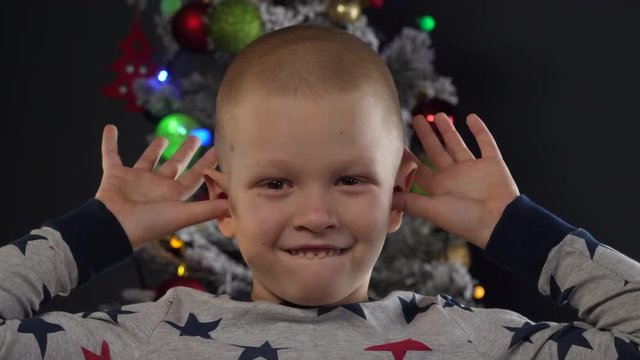 happy child shows grimaces in front of Christmas tree in garlands and green UFO balls of fire, glowing spheres, flashing lights, proton purple angels, plastic pink toys. New Year's Eve