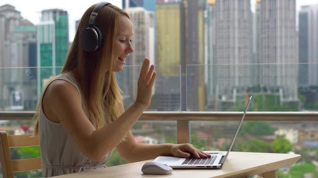 Young woman freelancer workes on her laptop at a balcony with a background of a city center full of skyscrapers. Remote techer has a conversation with a student through the internet