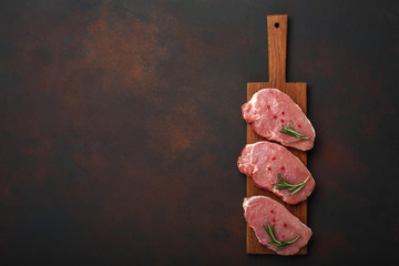 Pieces of raw pork steak on cutting board with rosemary and red pepper on rusty brown background
