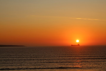 Orange sunset with silhouetted oil tanker on the sea and textured waves in the foreground.