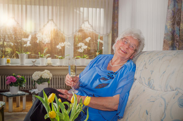 Seniorin entspannt sich auf eigenem Sofa und trinkt Sekt. Sie ist festlich gekleidet. Strauß gelbe...
