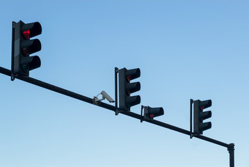 Outdoor surveillance camera and four red traffic lights installed on a pole above a roadway,...