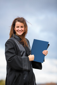 Pretty, young woman celebrating joyfully her graduation - spreading wide her arms, holding her diploma, savouring her success (color toned image; shallow DOF).
