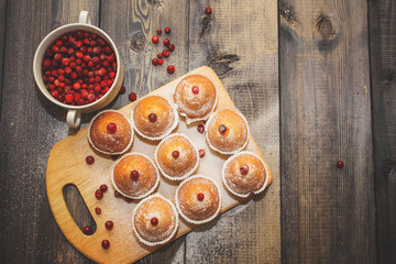 A cup with fresh red berries. Freshly baked muffins laid out on a wooden cutting board decorated with red berries on a light wooden table peppered with sugar.