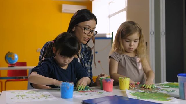 Cheerful mixed race female teacher in eyeglasses and two lovely preschool multicultural girls hand painting in kindergarten. Engrossed diverse kids keeping busy in hand painting activity at art lesson