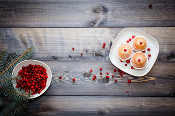 A light wooden table top with a plate of freshly baked muffins decorated with red berries sprinkled with white powder and a plate of red berries with spruce twigs.