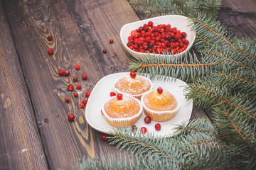 A light wooden table top with a plate of freshly baked muffins decorated with red berries sprinkled with white powder and a plate of red berries with spruce twigs.