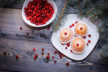 A light wooden table top with a plate of freshly baked muffins decorated with red berries sprinkled with white powder and a plate of red berries with spruce twigs.