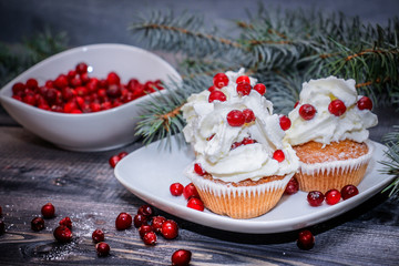 Freshly baked cake on a white plate decorated with red berries and whipped white cream with a plate topped with red berries decorated with spruce sprigs on a light wooden table top.