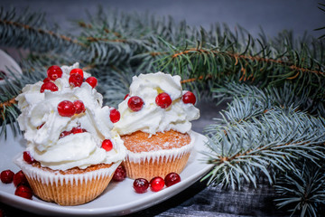 Freshly baked cake on a white plate decorated with red berries and whipped white cream with a plate topped with red berries decorated with spruce sprigs on a light wooden table top.