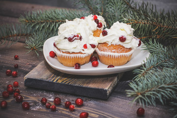 Freshly baked cake on a white plate decorated with red berries and whipped white cream with a plate topped with red berries decorated with spruce sprigs on a light wooden table top.