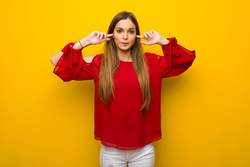 Young girl with red dress over yellow wall covering both ears with hands