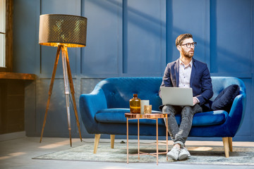 Portrait of an elegant businessman sitting with laptop on the couch at the luxury blue office interior