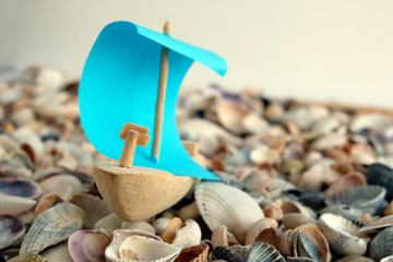 A toy ship with a blue sail is cast ashore with seashells and stones.