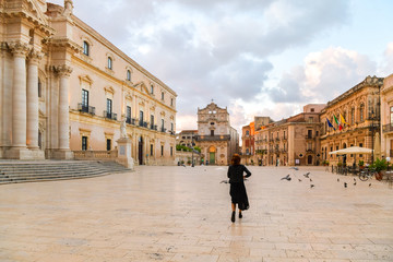 Fototapeta na wymiar Young woman is running across the main square with pigeons, island of Ortigia, Syracuse, Italy