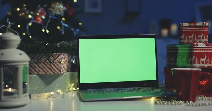 Close up of the laptop computer with green screen being on the table in the dark room surrounded by Christmas presents and Christmas tree. Alpha channel. Chroma key. Inside.