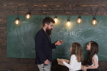Teacher and girls pupils in classroom near chalkboard. Man with beard in formal suit teaches...