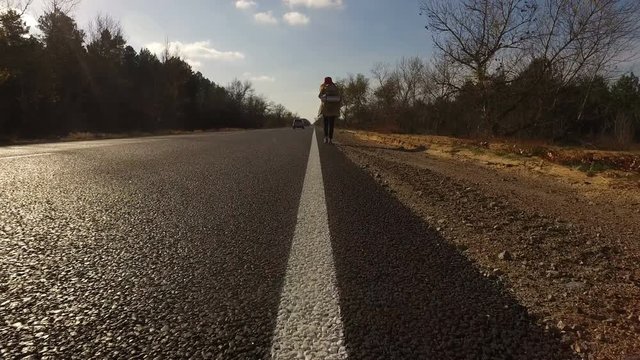 Young girl, tourist hitchhiker walks along road with a backpack on his shoulders, lifting his thumb up. Traveler is trying to stop car. Highway in summer forest