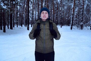 A young man in a green jacket and a hat with a backpack stands on the background of a snowy pine forest. Man and winter.