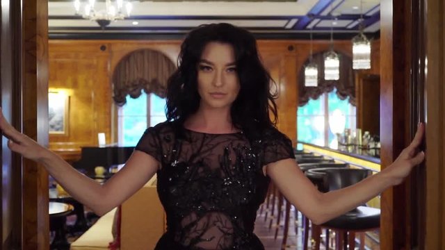 Young sexy brunette woman in black evening dress opens the doors and going out. Slowmotion. In bar, restaurant or hotel lobby. Luxury