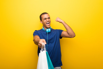 African american man with blue t-shirt on yellow background holding a lot of shopping bags