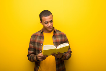 Young african american man on vibrant yellow background holding a book and enjoying reading