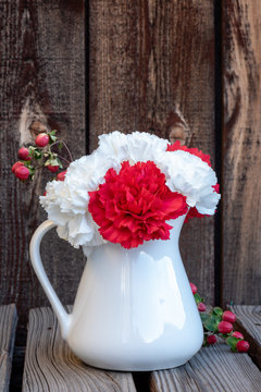 Close up of a  white pitcher with white and red carnations and a stem of red berries on a rustic plank table and plank background.
