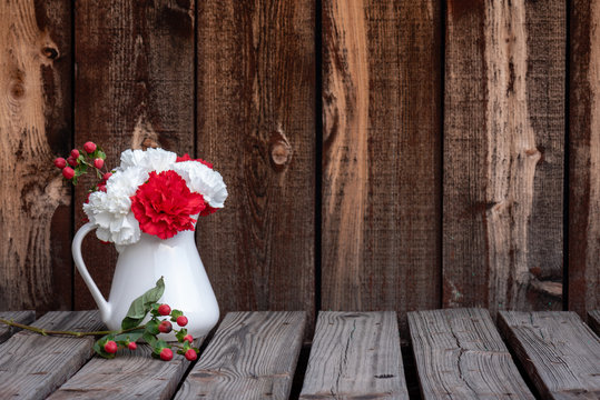 White pitcher with white and red carnations and a stem of red berries on a rustic plank table and plank background.
