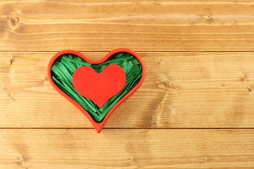 Red painted wooden heart enclosed with green paper raffia strips in red box isolated on wooden background