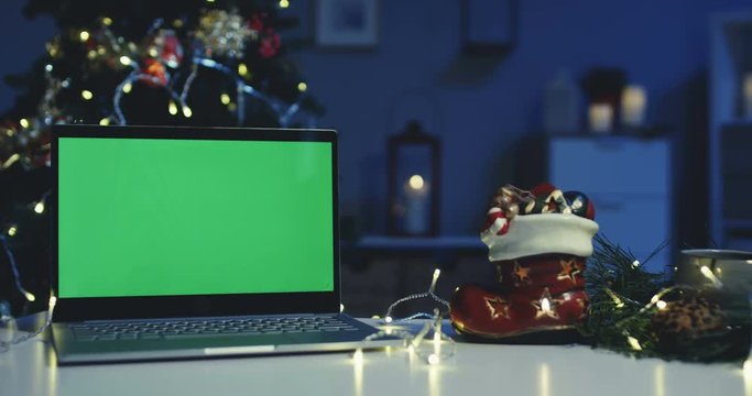 Laptop computer with a green screen at the Christmas tree on the table at the Christmas boot with presents at cozy home in the evening. Close up. Indoor.
