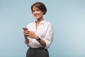 Young pretty smiling woman with dark short hair in white shirt happily using cellphone over blue...