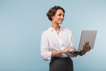 Young smiling businesswoman with dark short hair in white shirt happily working on laptop over blue...