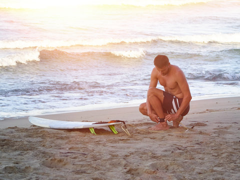 young man on the beach, fastens the leash to the foot from the surfboard, preparing to surf during sunset