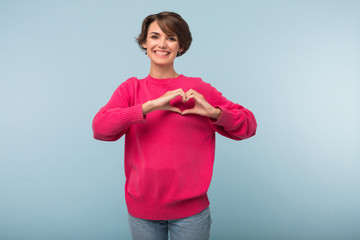 Beautiful smiling girl with dark short hair in pink sweater showing heart gesture with hands while...