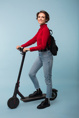 Young pretty smiling girl in red sweater and jeans with black backpack standing with electric scooter while happily looking in camera over blue background