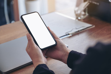 Mockup image of hands holding black mobile phone with blank white screen with laptop on wooden table